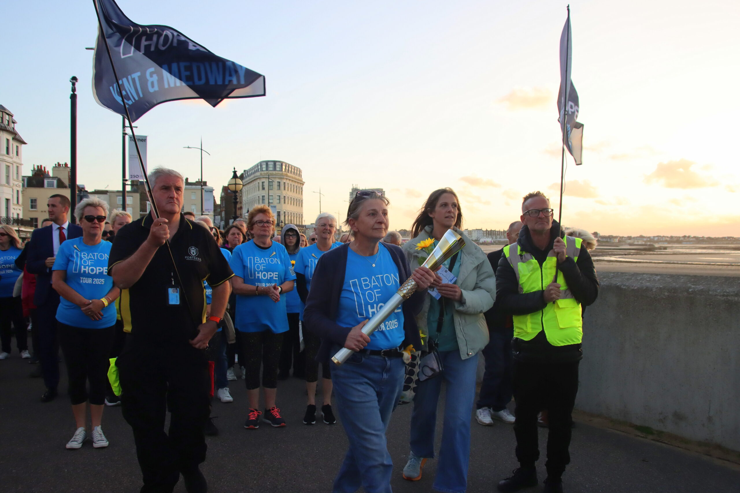 A group of people walking alongside a beach in Kent with flags and one person holding an elaborate metal baton.