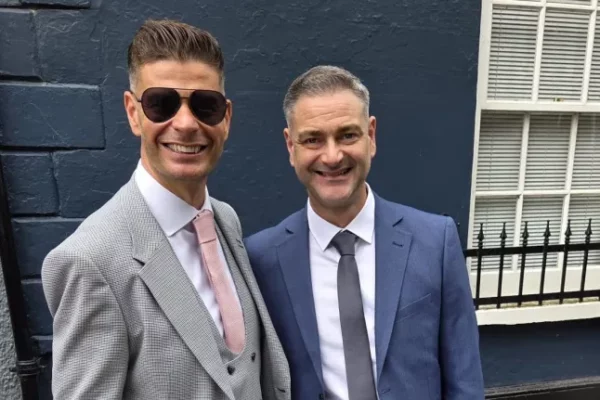 A photo of two men stoof in front of a brick wall wearing suits and smiling.
