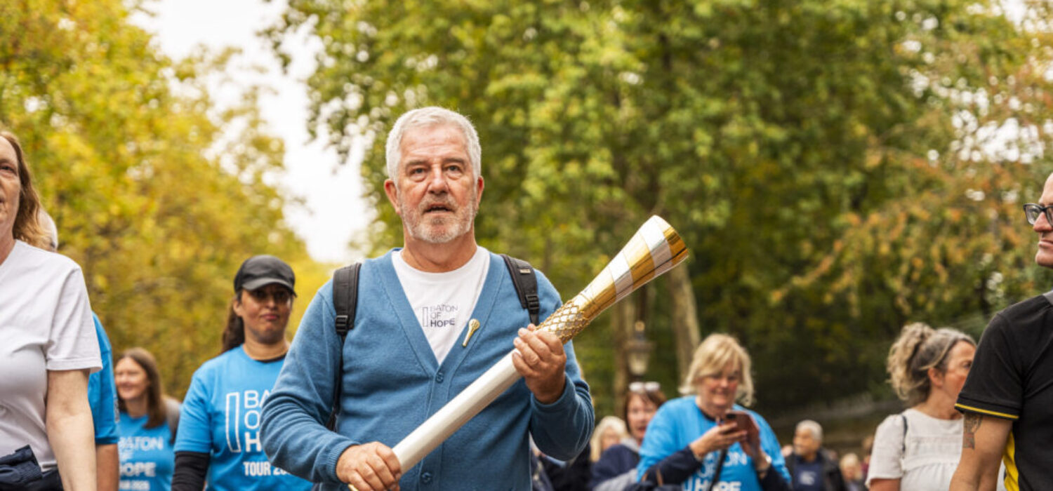 A man walking outside in a park holding an elaborate metal baton with people walking behind him.
