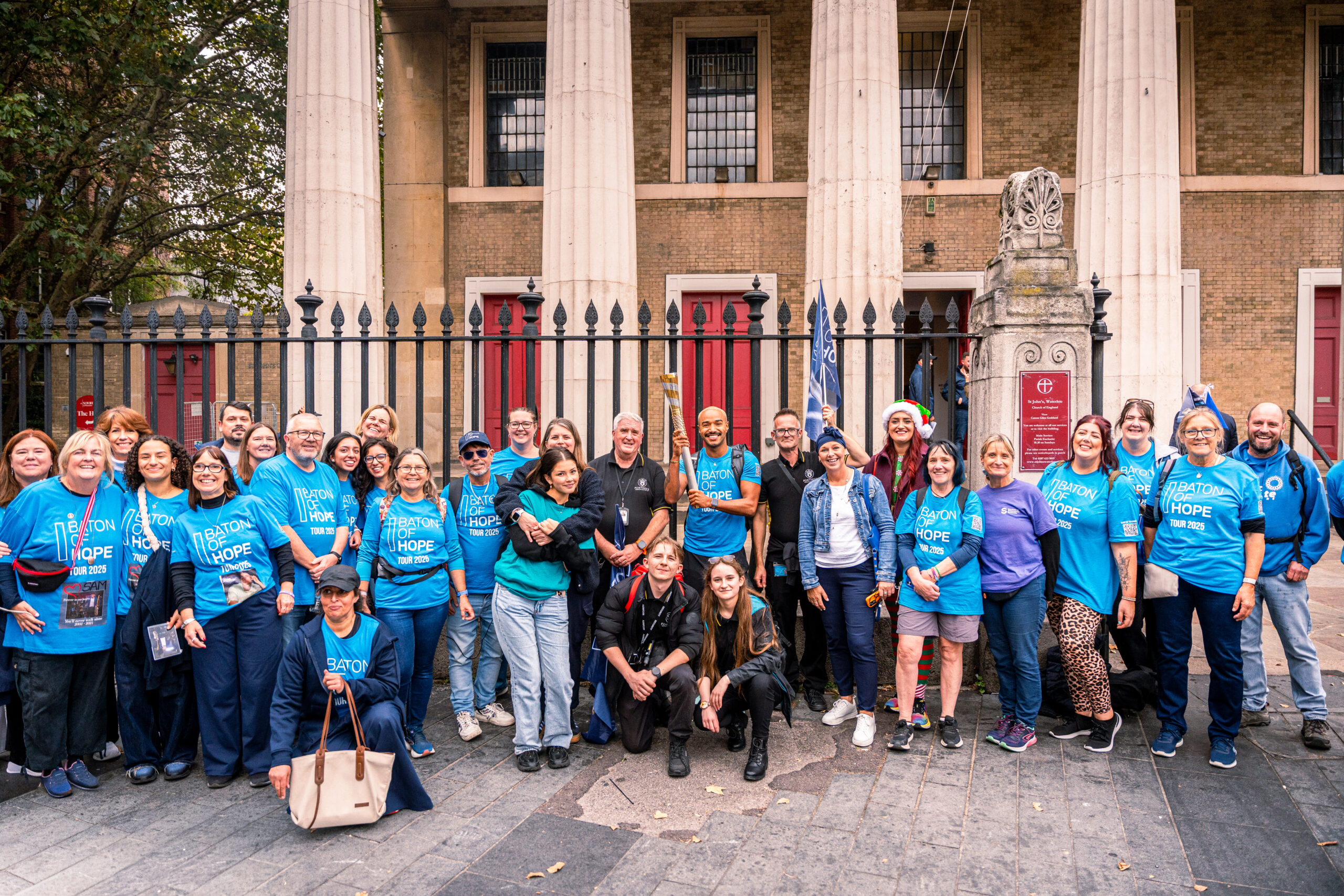 A group of people in light blue t-shirts stood as a group outside in front of a building.
