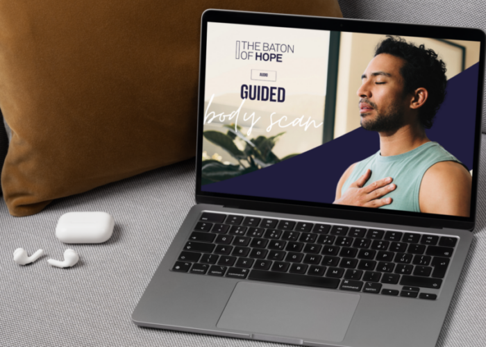 A mockup design showing a laptop on a safe with a man on the computer screen with his eyes closed and a hand on his chest in a room with the text "The Baton of Hope Audio Guided Body Scan" on the image.